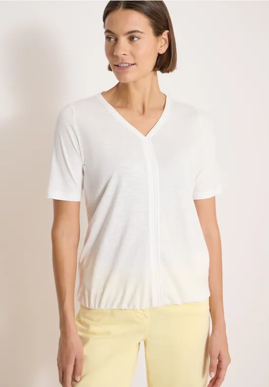 Een vrouw draagt een wit V-hals T-shirt en een gele broek tegen een effen achtergrond.