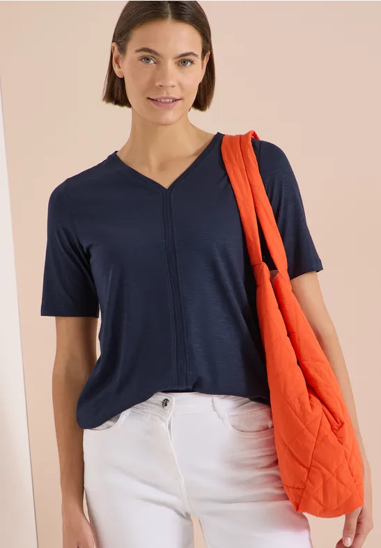 Eine Frau trägt ein dunkelblaues V-Shirt, weiße Hose und eine orange Stepp-Shopper-Tasche.