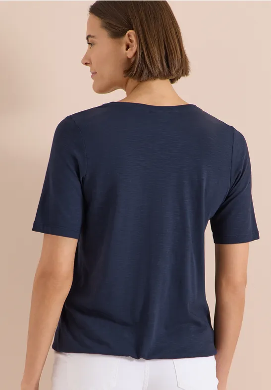 Rückansicht einer Person mit schulterlangem braunem Haar und dunkelblauem T-Shirt vor hellem Hintergrund.