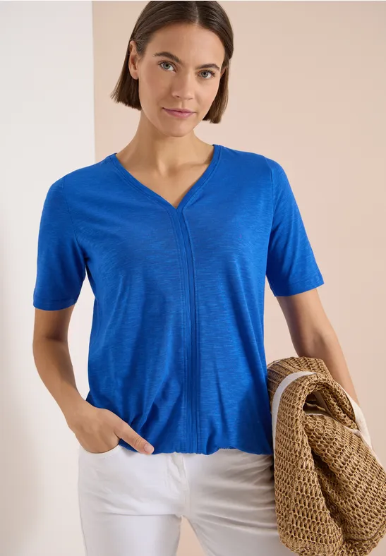 Femme portant un t-shirt bleu à col V et un pantalon blanc, tenant un panier tressé.