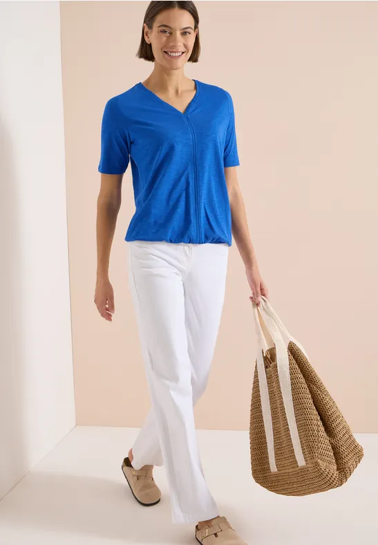 Femme en haut bleu col V, pantalon blanc et chaussures beiges, portant un grand sac en osier.