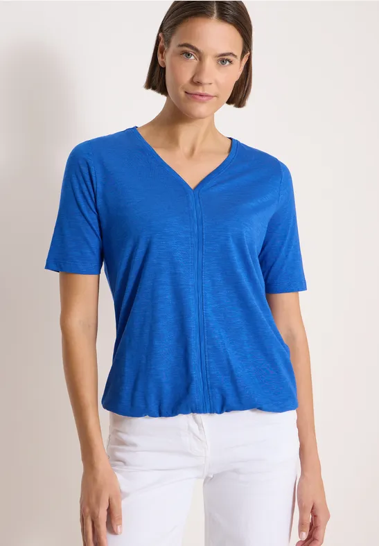 Femme porte un t-shirt bleu vif à col en V et un pantalon blanc, sur fond clair.