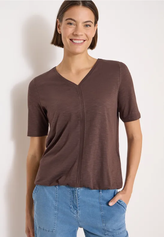 Een persoon draagt een bruin V-hals t-shirt met korte mouwen en steekt een hand in een lichtblauwe spijkerbroek.