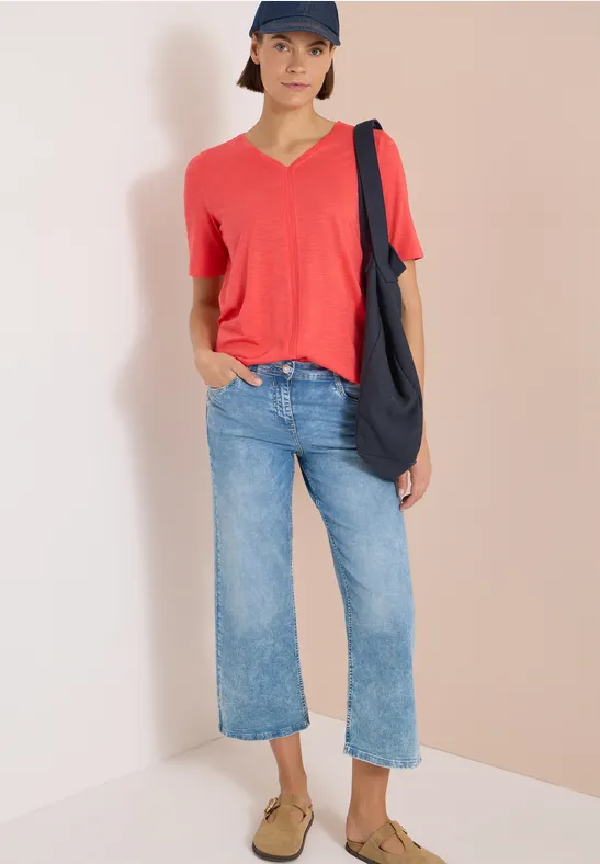 Person trägt rotes V-Shirt, weite blaue Jeans, beigen Slipper und dunkle Tasche vor hellem Hintergrund.