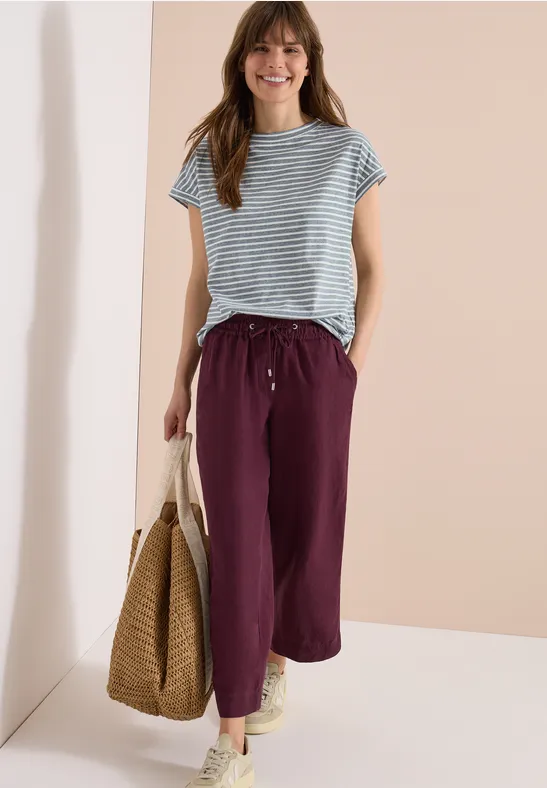 Een vrouw met een gestreepte top, paarse broek en een strooien tas.