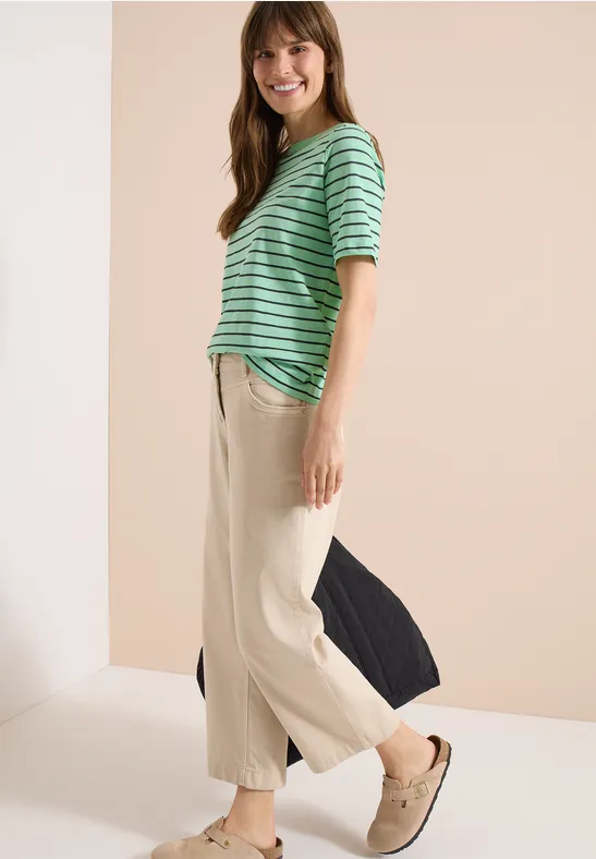 Frau im Profil trägt gestreiftes grünes Oberteil, helle weite Hosen und Slip-ons vor neutralem Hintergrund.