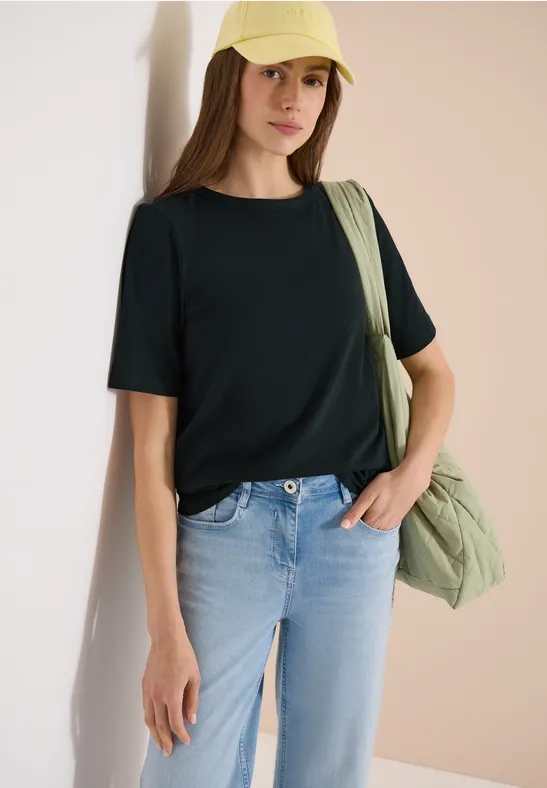 Frau trägt schwarzes T-Shirt, helle Jeans und eine grüne Umhängetasche vor neutralem Hintergrund.