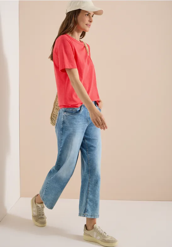 Frau im roten T-Shirt, blauen Jeans und Sneakers, seitlich stehend vor beigem Hintergrund.