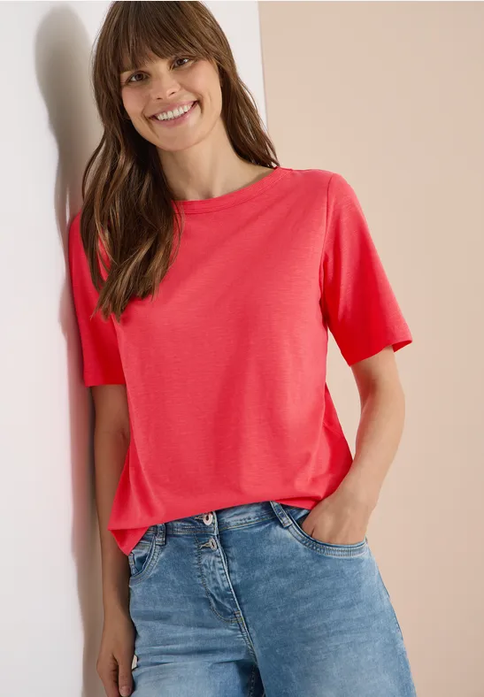 Frau in rotem T-Shirt und blauer Jeans lehnt lässig an der Wand, eine Hand in der Hosentasche