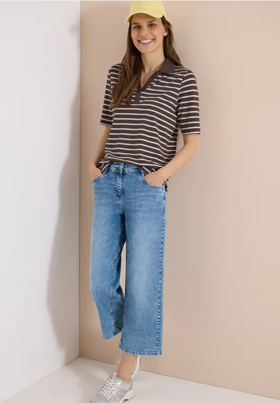 Personne en polo rayé, jean large et baskets blanches posant décontractée contre un mur clair.