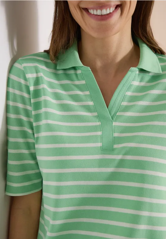 Eine Frau lehnt lächelnd an einer Wand und trägt ein grün-weiß gestreiftes Poloshirt.