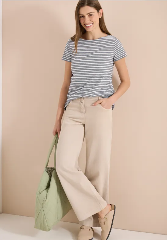 Frau in gestreiftem T-Shirt, weiten beigen Hosen, grüner Tasche und beigen Slippern vor neutralem Hintergrund