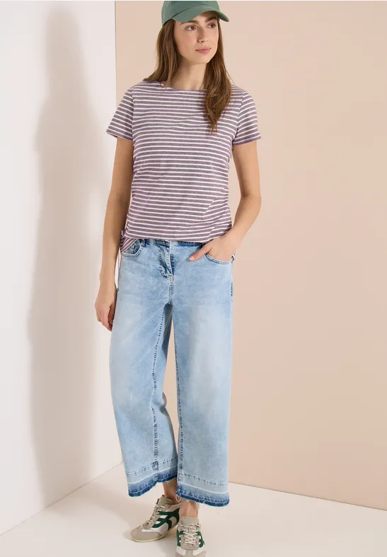 Person mit gestreiftem T-Shirt, weitem Jeans und Sneakern steht in einem hellen Eckzimmer.