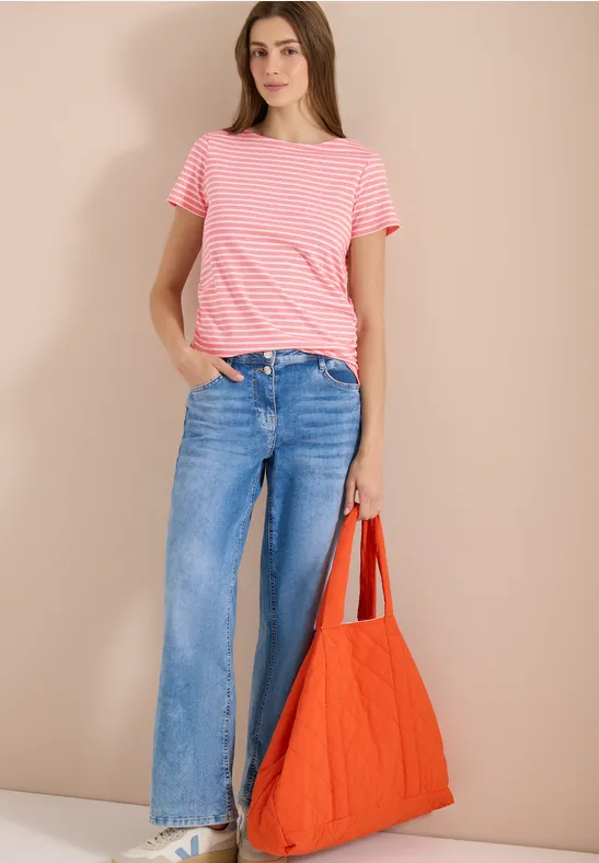 Frau in rosa gestreiftem T-Shirt, weiter Jeans und roter Tasche vor neutralem Hintergrund.