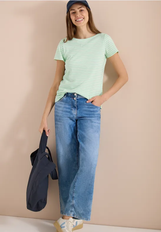 Personne devant un fond beige, porte un t-shirt vert rayé, jean large, baskets blanches et un sac noir.