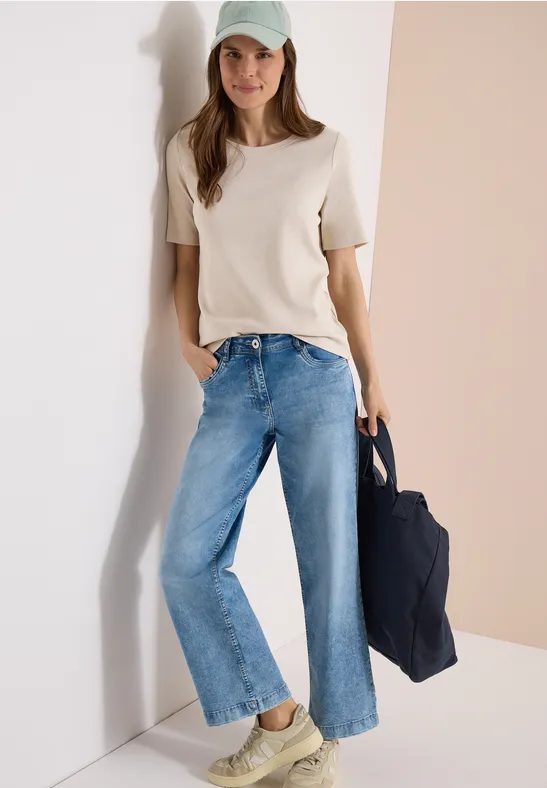 Frau in weißem T-Shirt und weitem Jeans, trägt Mütze und Tasche, lehnt elegant an heller Wand.