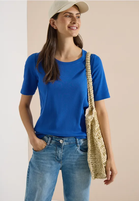 Vrouw in blauw T-shirt en spijkerbroek, met stro tas en pet, staat nonchalant met één hand in de zak
