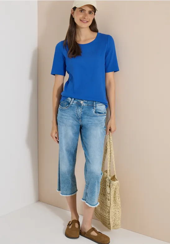 Vrouw in blauw T-shirt, caprijspantalon, strooien tas en bruine sloffen voor een neutrale muur.