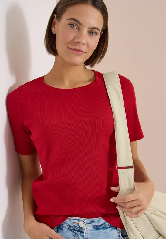 Person lehnt an Wand, trägt rotes T-Shirt, blaue Jeans und eine helle Schultertasche.