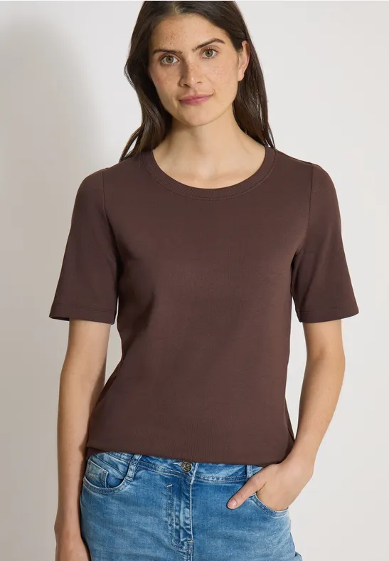 Vrouw draagt bruin T-shirt met korte mouwen en blauwe spijkerbroek, hand in de zak.
