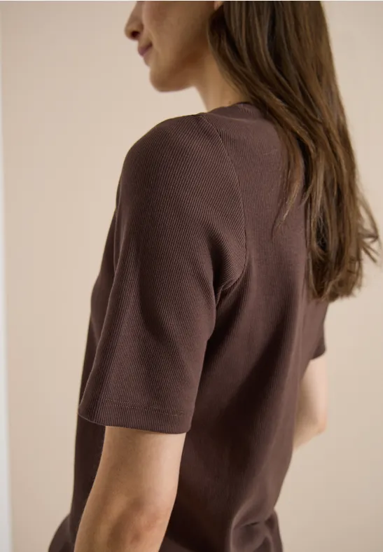 Vrouw van achteren in een bruin t-shirt, lange donkere haar, neutrale achtergrond.
