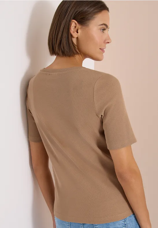 Frau mit kurzem Bob von hinten, trägt beige Kurzarmshirt, lehnt an weißer Wand.