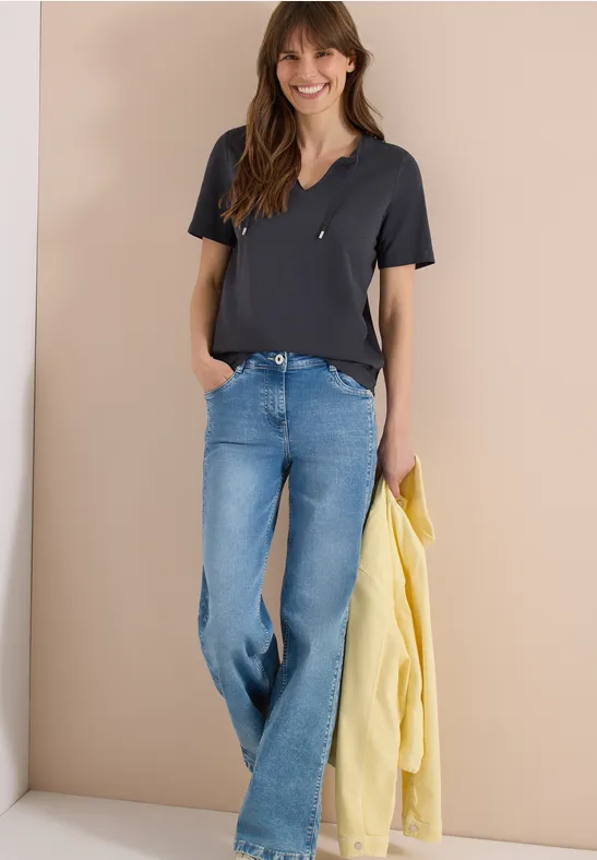 Frau in schwarzem T-Shirt und blauen Jeans hält eine gelbe Jacke vor beigem Hintergrund.