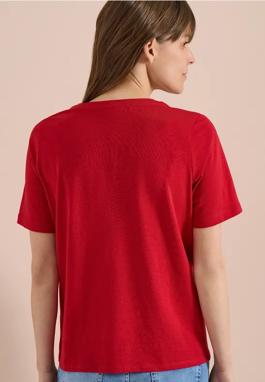 Frau von hinten in rotem Kurzarmshirt, Jeans und weißen Sneakern vor neutralem Hintergrund.