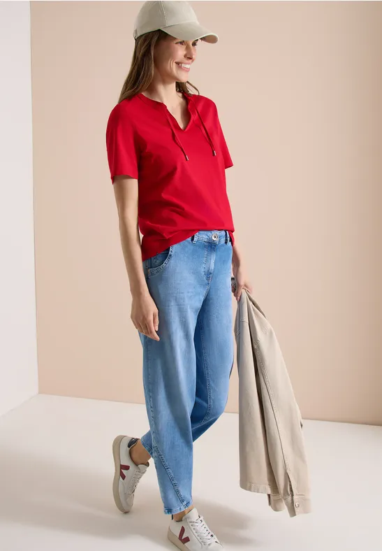 Frau in rotem Poloshirt, blauen Jeans und Cap, hält beigen Mantel in hellem Umfeld entspannt.