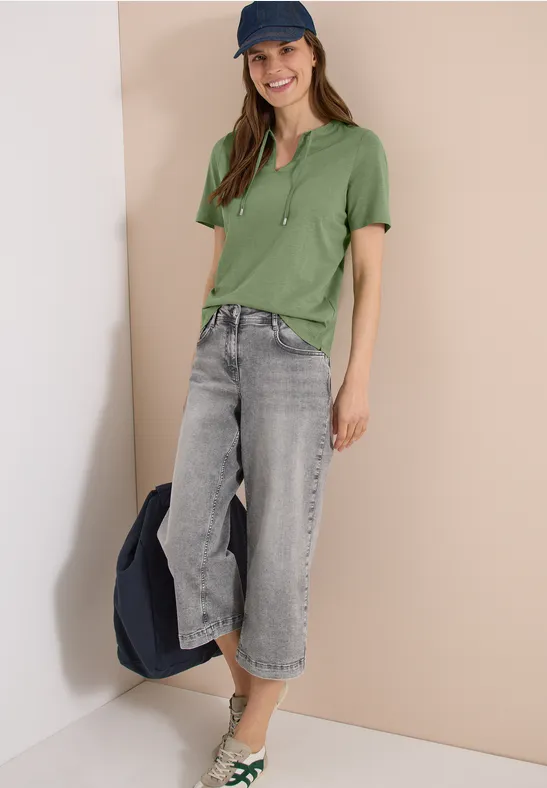 Une personne porte un polo vert, un jean gris ample et une casquette bleu marine, tenant une veste.