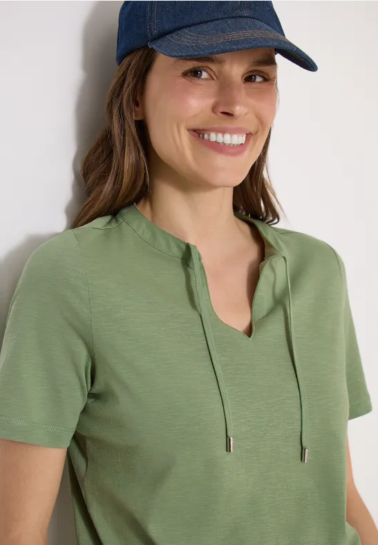 Femme en t-shirt vert à manches courtes avec cordons au col en V et casquette bleue.