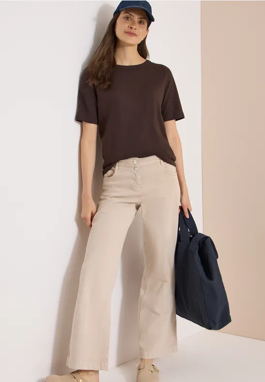 Vrouw leunt tegen een muur, draagt een donkerbruin T-shirt, wijde beige broek en houdt een donkerblauwe tas.