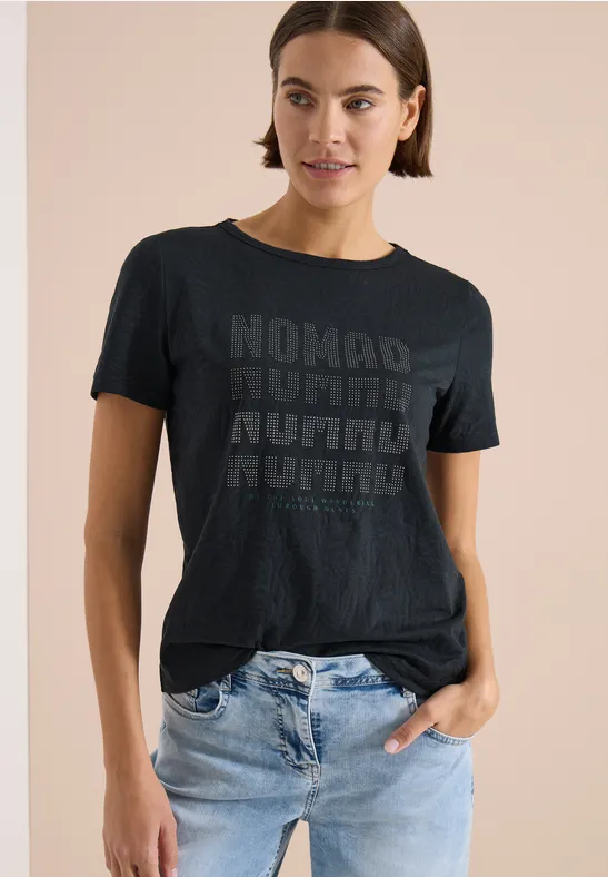 Eine Person trägt ein schwarzes T-Shirt mit wiederholtem 'NOMAD'-Schriftzug und helle Jeans.