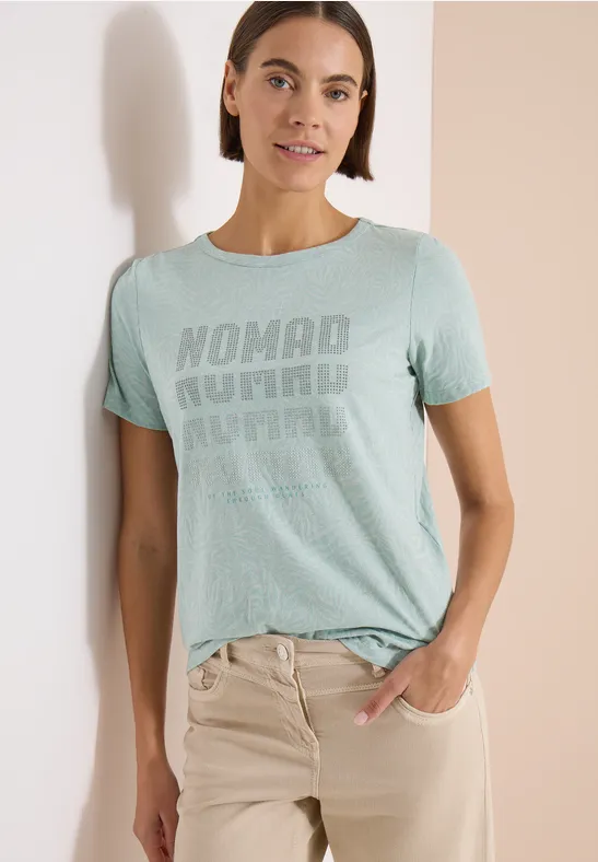 Eine Frau trägt ein mintgrünes T-Shirt mit 'Nomad Journal'-Print und beigen Hosen.