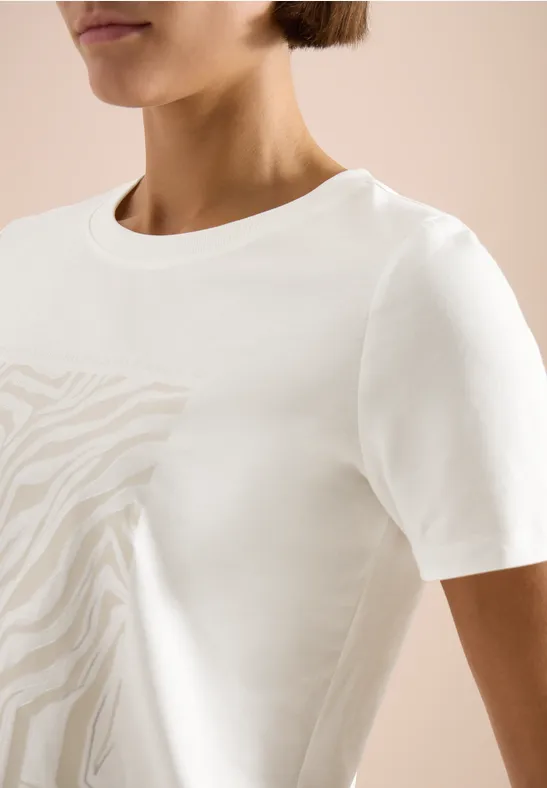 Frau trägt ein weißes T-Shirt mit dezentem Zebramuster vor beigem Hintergrund.
