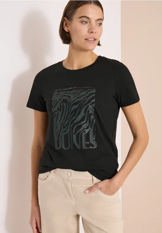Frau im schwarzen T-Shirt mit grafischem Muster und beiger Hose vor hellem Hintergrund.