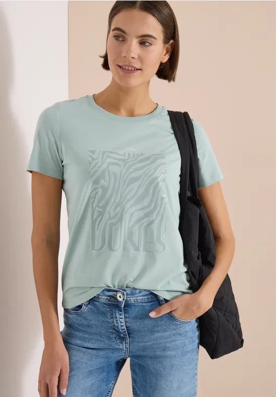 Frau im hellgrünen T-Shirt mit Muster, blauer Jeans und schwarzer Tasche, eine Hand in der Tasche.