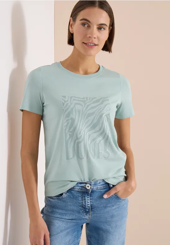 Person trägt hellgrünes T-Shirt mit abstraktem Zebramuster und Jeans vor hellem Hintergrund.