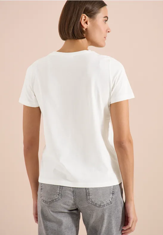 Frau steht vor hellem Hintergrund, trägt weißes T-Shirt und graue Jeans, Rückenansicht.