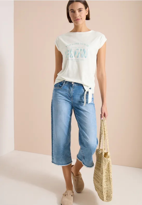 Vrouw met wit T-shirt, wijde culotte-jeans, strooien tas en espadrilles tegen een lichte achtergrond.
