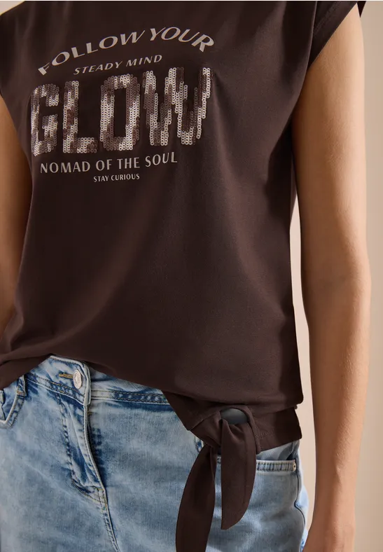 Dunkelbraunes T-Shirt mit Schriftzug, seitlicher Schlitz und Knoten, kombiniert mit hellblauen Jeans.