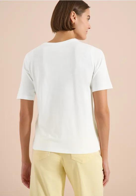 Person von hinten mit kurzem braunen Haar, weißem T-Shirt und gelber Hose vor beigem Hintergrund.
