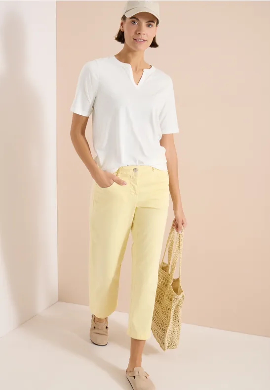 Frau trägt weißes V-Shirt, helle gelbe Hose und beige Sneaker, hält geflochtene Strohtasche vor hellem Hintergrund.