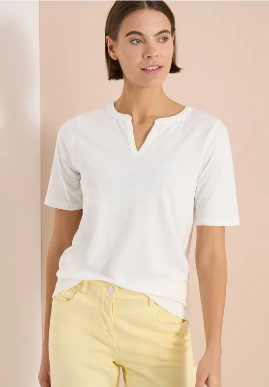 Eine Frau trägt ein weißes V-Ausschnitt-Shirt und hellgelbe Jeans vor neutralem Hintergrund.
