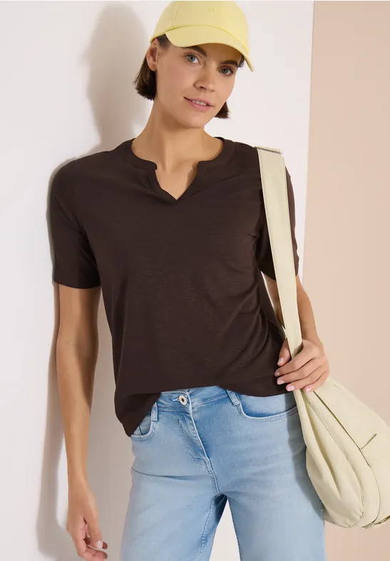 Frau in dunklem T-Shirt, hellblauen Jeans, hellbeigem Schulterbeutel und Cap vor zweifarbiger Wand
