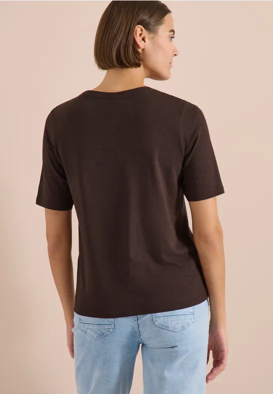 Frau von hinten in braunem T-Shirt mit Halbarm und hellblauer Jeans vor beigem Hintergrund.