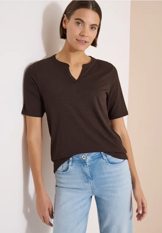 Eine Person in dunklem T-Shirt mit V-Ausschnitt und hellen Jeans vor hellem Hintergrund.