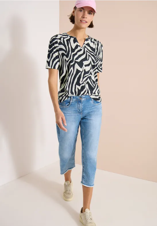 Persoon draagt zwart-wit zebramotief-shirt, korte blauwe jeans en sneakers tegen neutrale achtergrond.