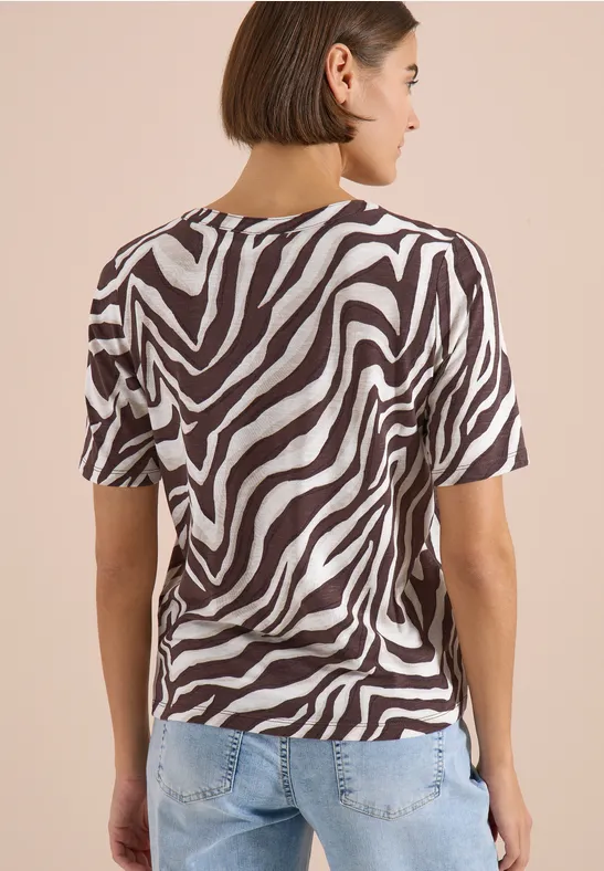 Iemand van achteren in een zwart-wit zebraprint t-shirt en een lichtblauwe spijkerbroek voor een beige achtergrond.
