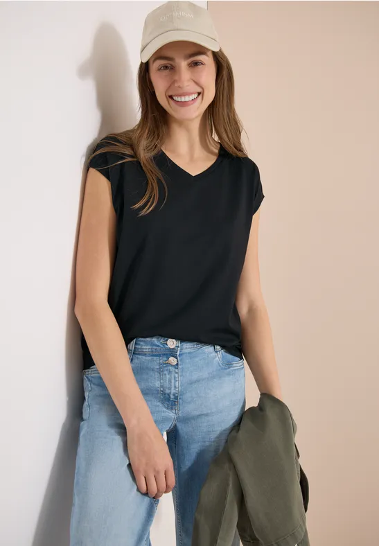 Une femme porte un T-shirt noir à col V manches courtes, jean bleu et tient une veste kaki à la main.